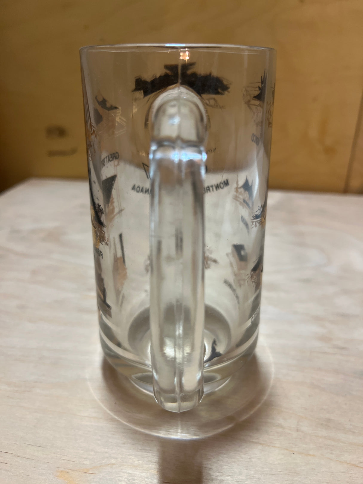 '67 Montreal Expo with National Pavilions souvenir mug, featuring printed illustrations of landmarks, sits empty on a light wooden surface with the handle facing forward.