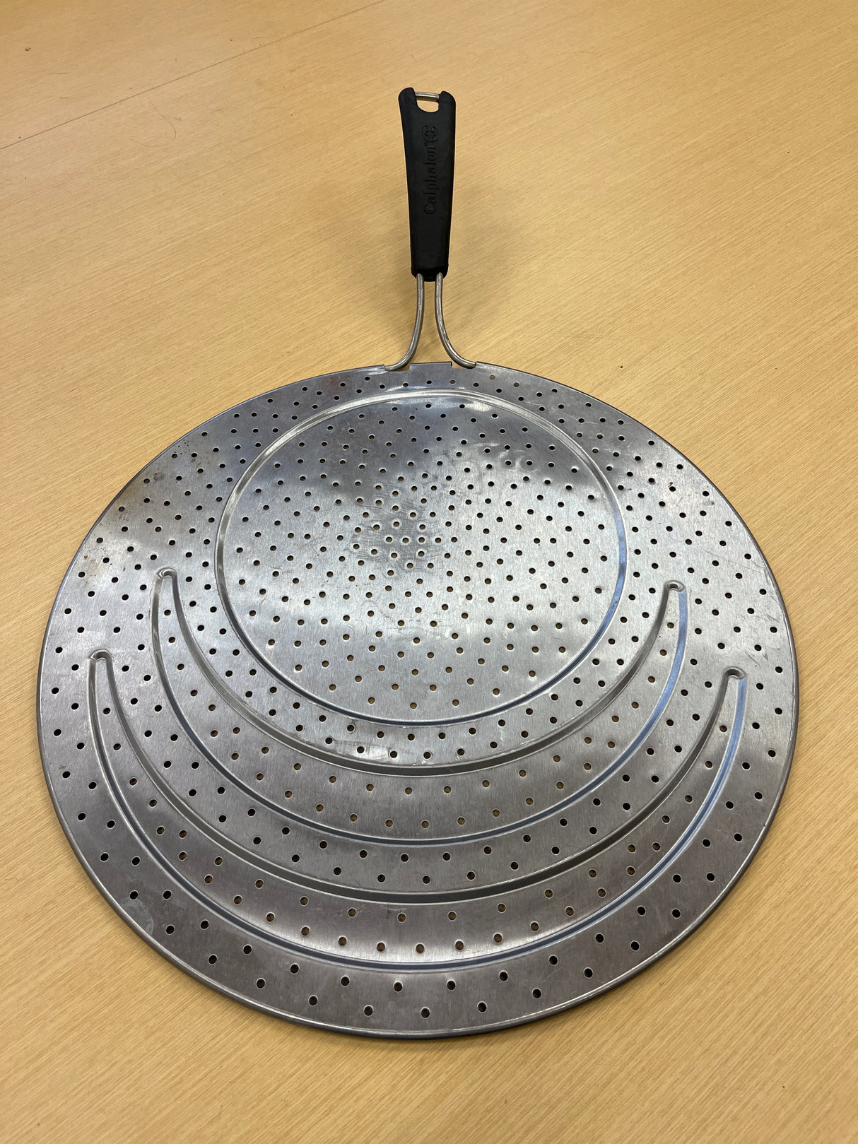 Calphalon 13 stainless steel splatter guard with silicone lined handle, featuring a perforated metal surface, designed to fit various pan sizes and keep stovetops clean.