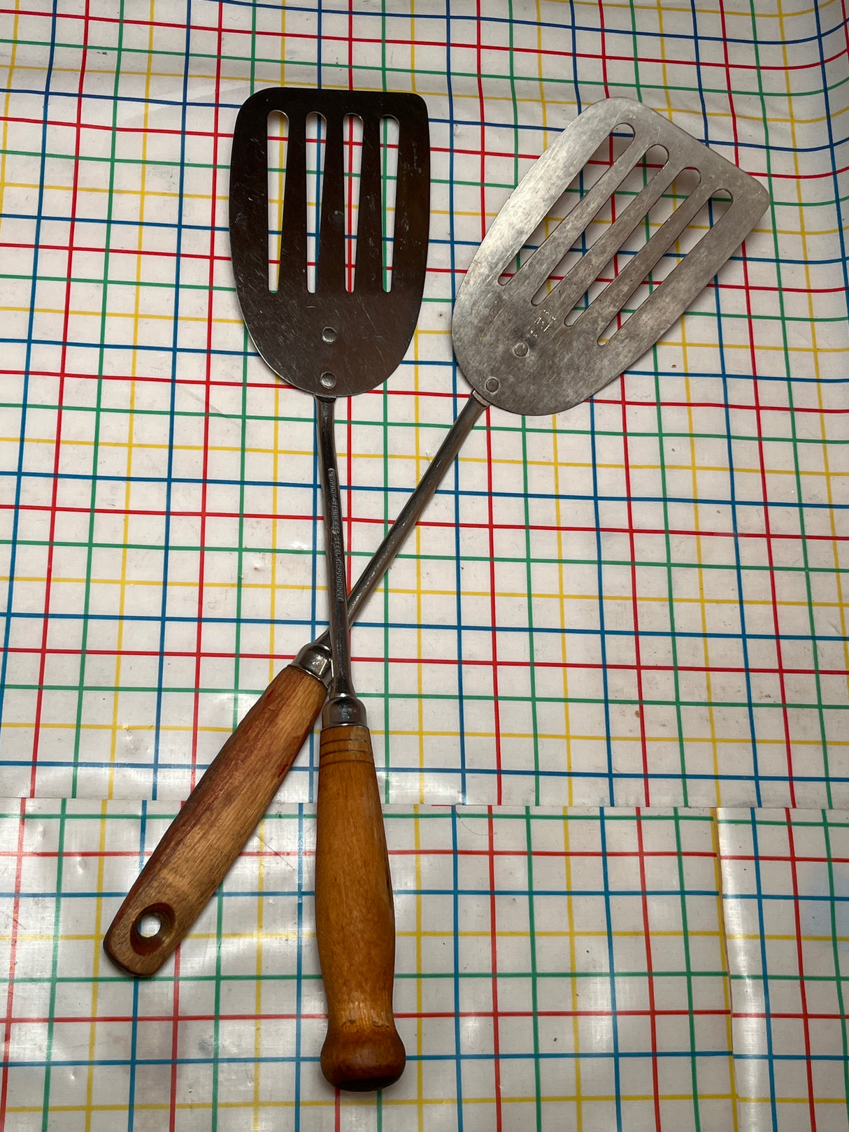 Two crossed vintage flat thin steel slotted spatulas with wood handles, ideal for flipping pancakes and grilling, on a patterned tablecloth.