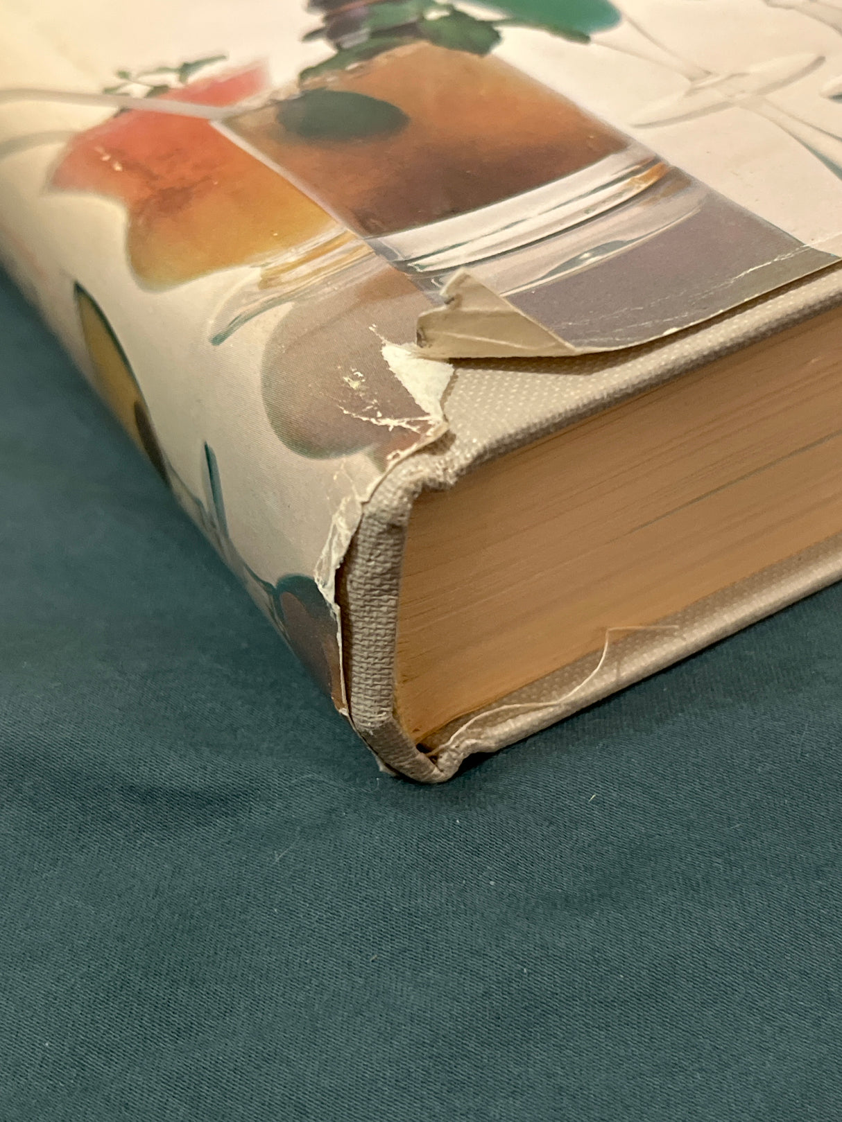 Trader Vic's Bartender's Guide (Revised 1972) showing a worn dust jacket corner, revealing cloth binding and page block, on a teal fabric surface.