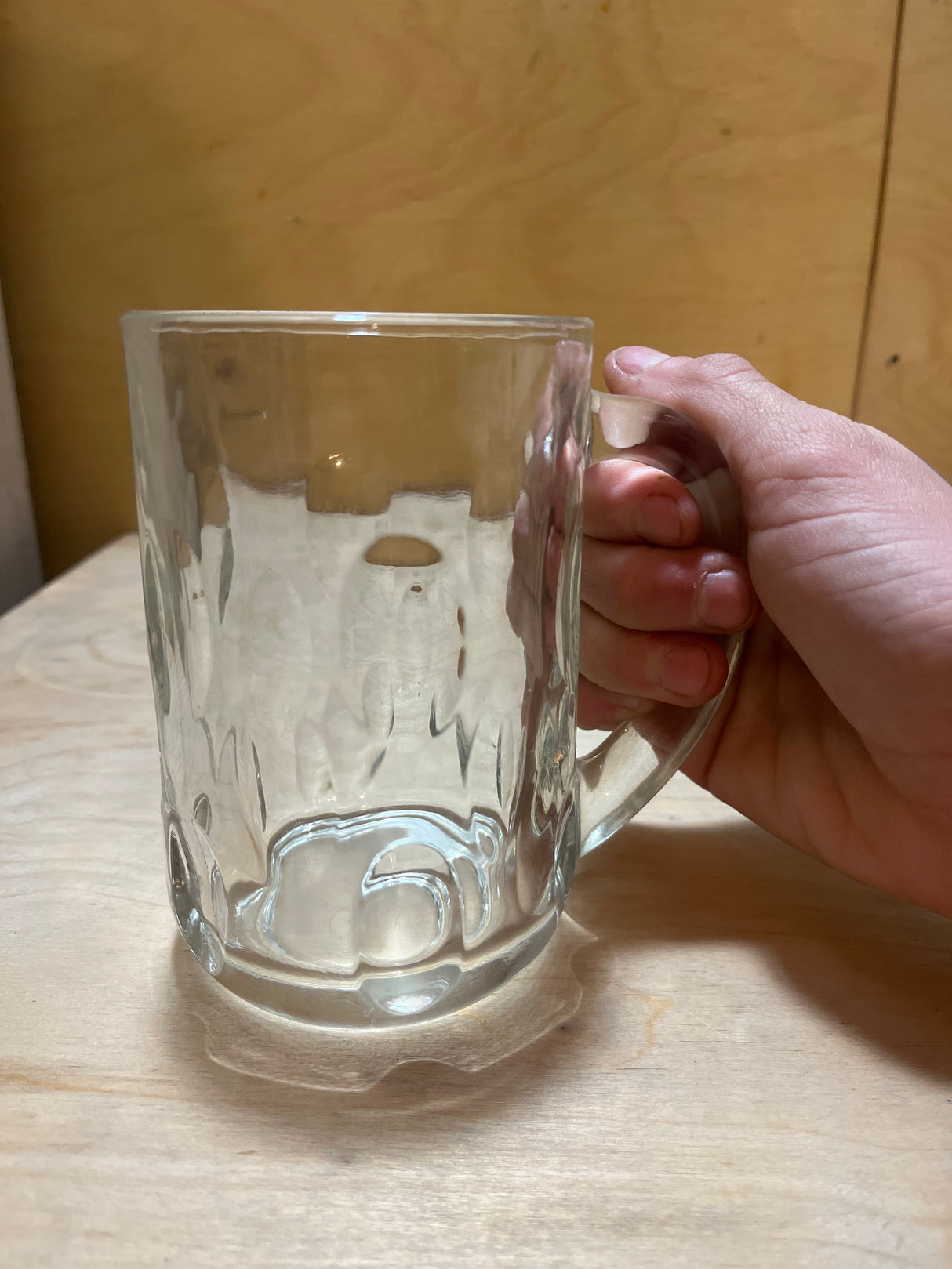 A right hand holds an Anchor Hocking vintage clear thumbprint dimple glass mug, showcasing its thick-walled design and sturdy handle on a wooden surface.