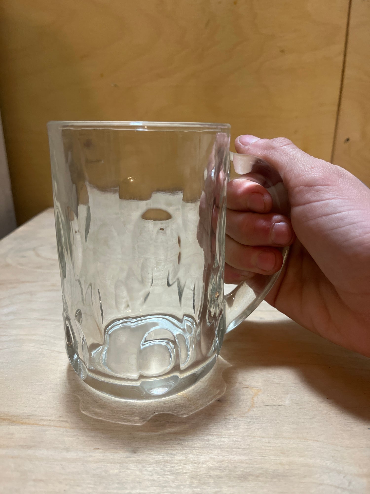 A right hand holds an Anchor Hocking vintage clear thumbprint dimple glass mug, showcasing its thick-walled design and sturdy handle on a wooden surface.
