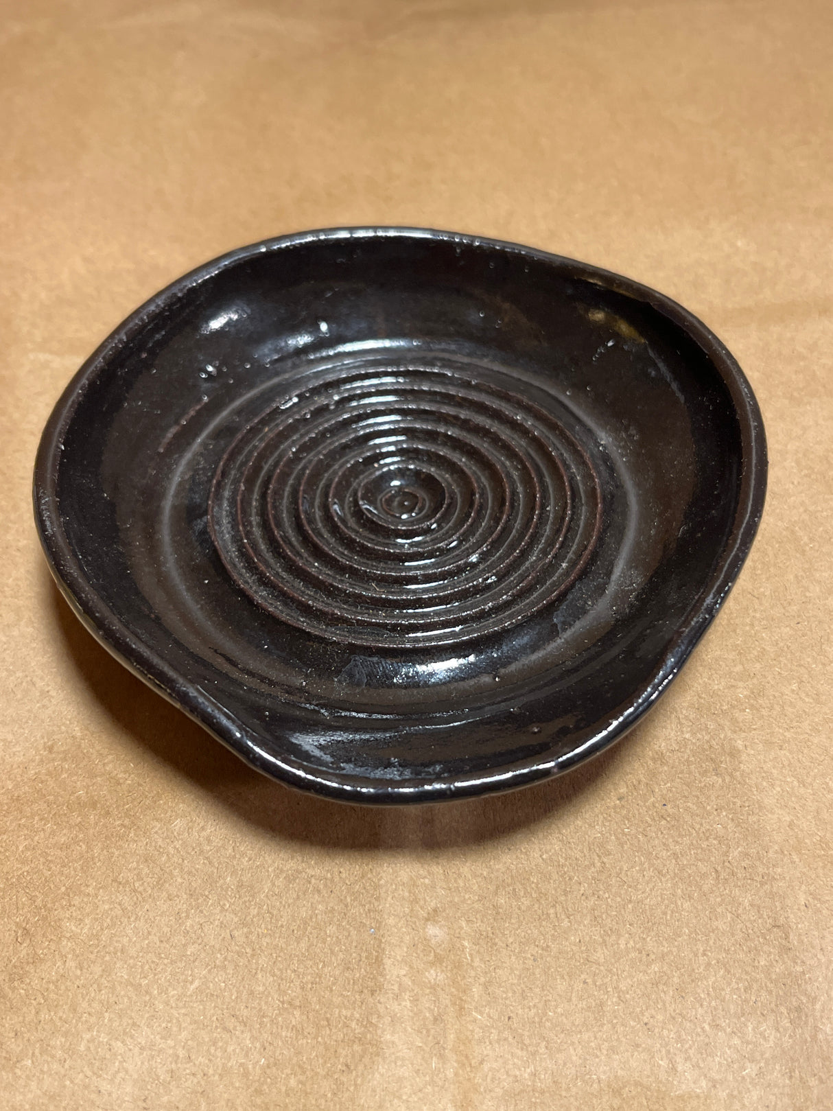 Handmade ceramic garlic grater spoon rest by Charles Lehman, featuring a glossy black glaze with concentric rings, set on cardboard.