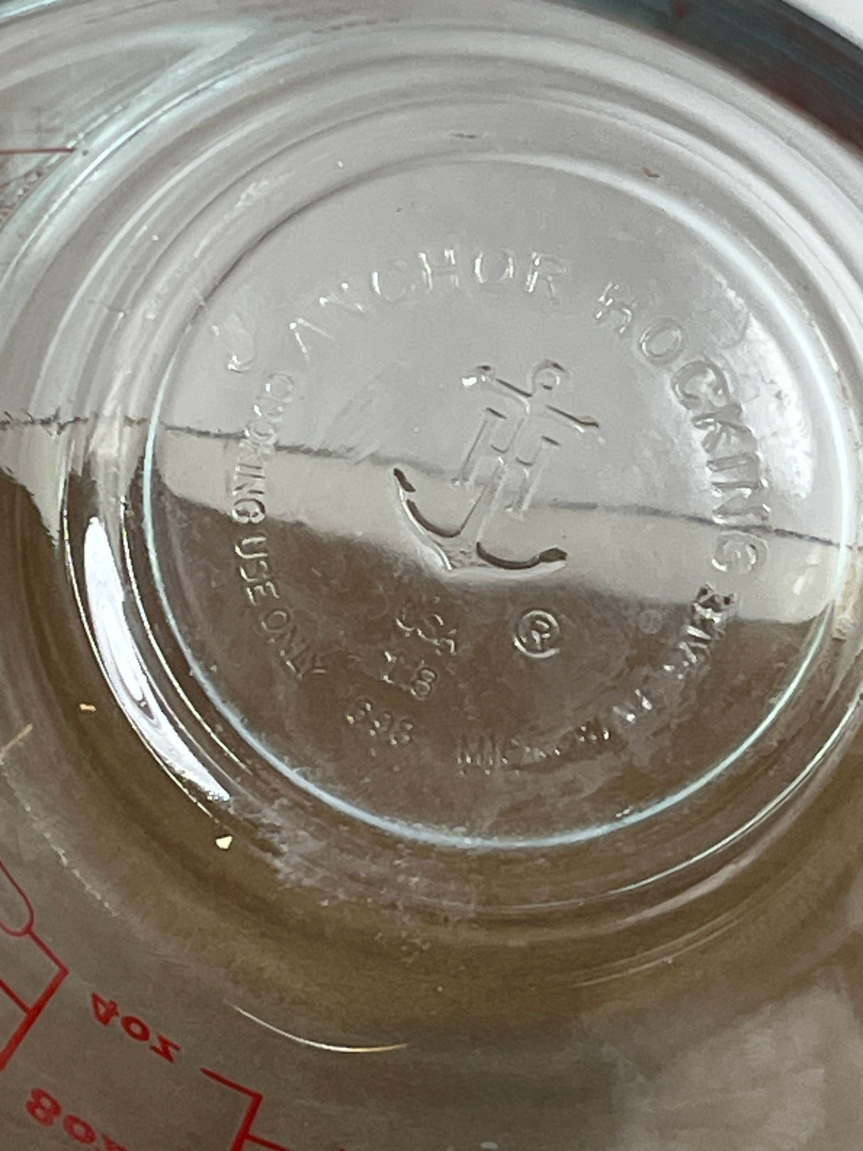 Underside view of an Anchor Hocking glass 2-cup measuring cup with handle, showing embossed anchor logo, safety markings, and partial red ounce-markings.