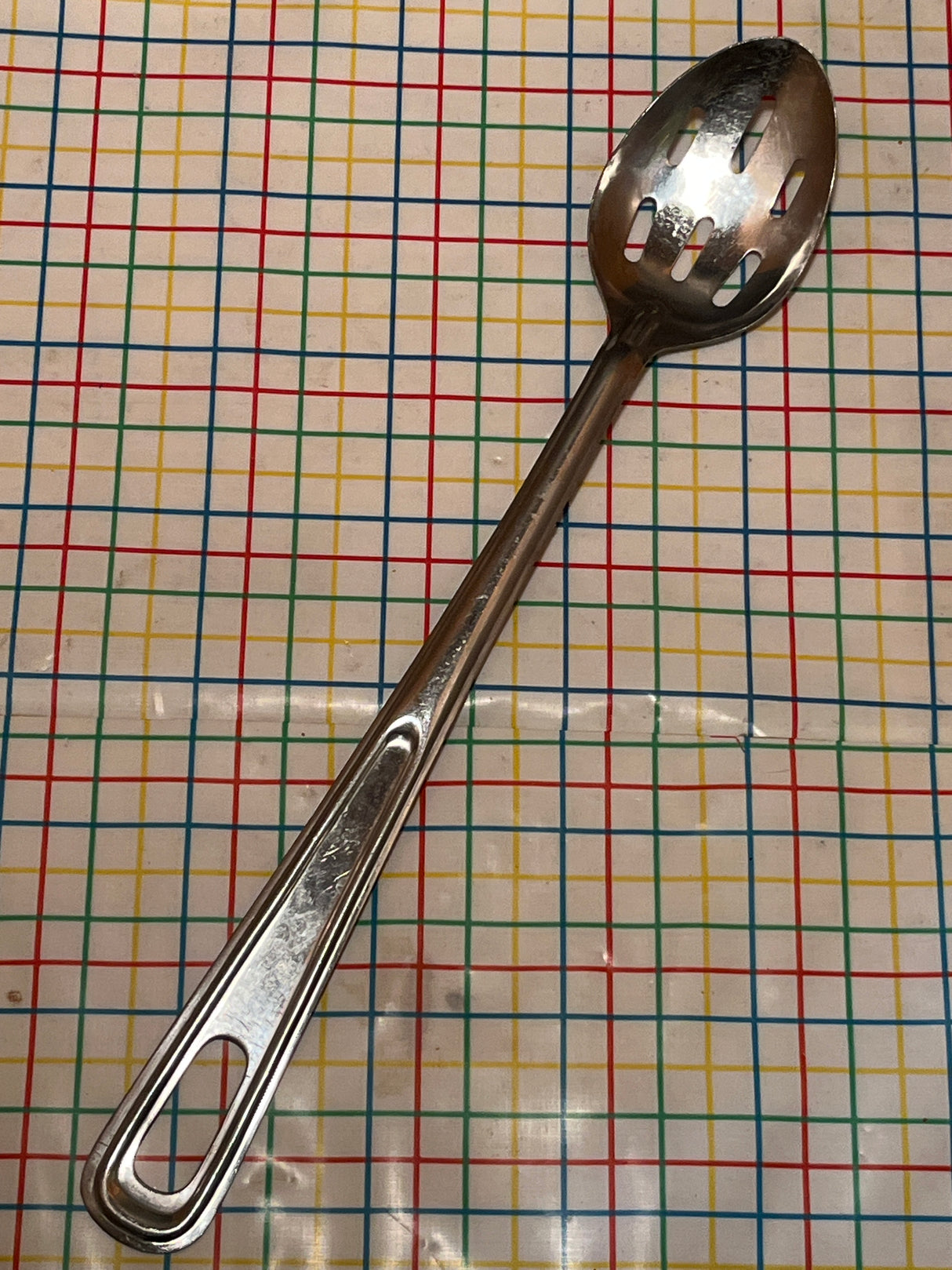 Extra long slotted basting spoon - JR stainless steel, featuring a durable, easy-to-clean long handle, resting on a tabletop with a grid-patterned tablecloth.