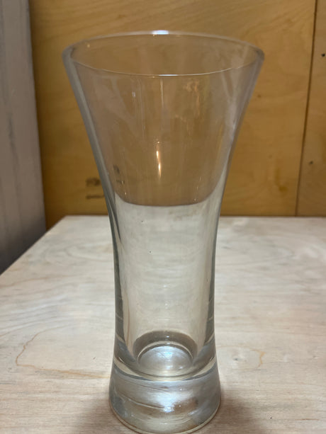 Tall Pilsner beer glass with a flared rim and thick rounded base, sitting empty on a light-wood surface against a plywood background.