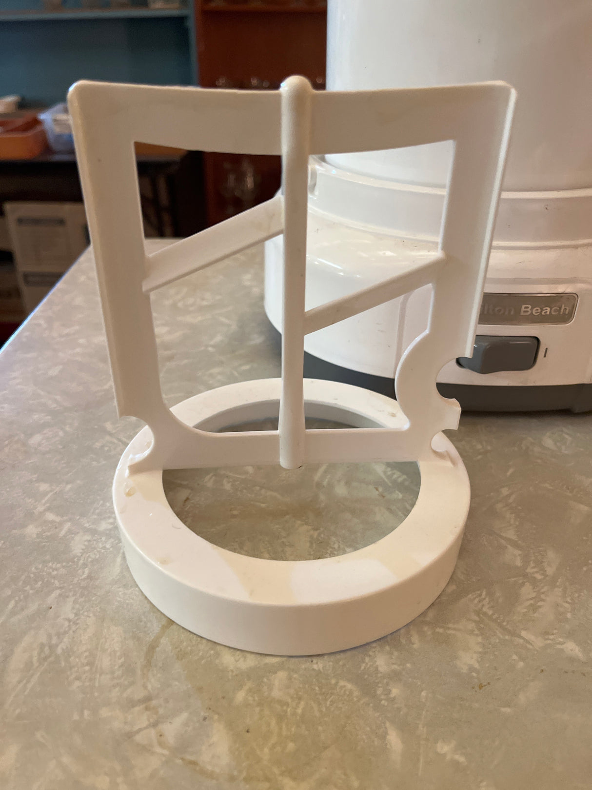 Automatic ice cream maker - Hamilton Beach 68880, featuring a plastic filter-basket bracket, part of a juicer attachment, seen with the main appliance in the background.