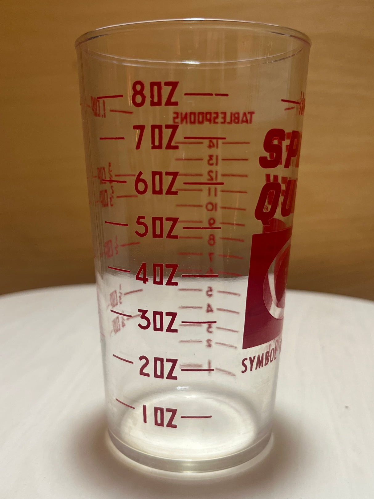 Speed Queen glass measuring cup with red gradations from 1 oz to 8 oz and tablespoon markings, sitting empty on a light-colored surface against a wood-grain backdrop.