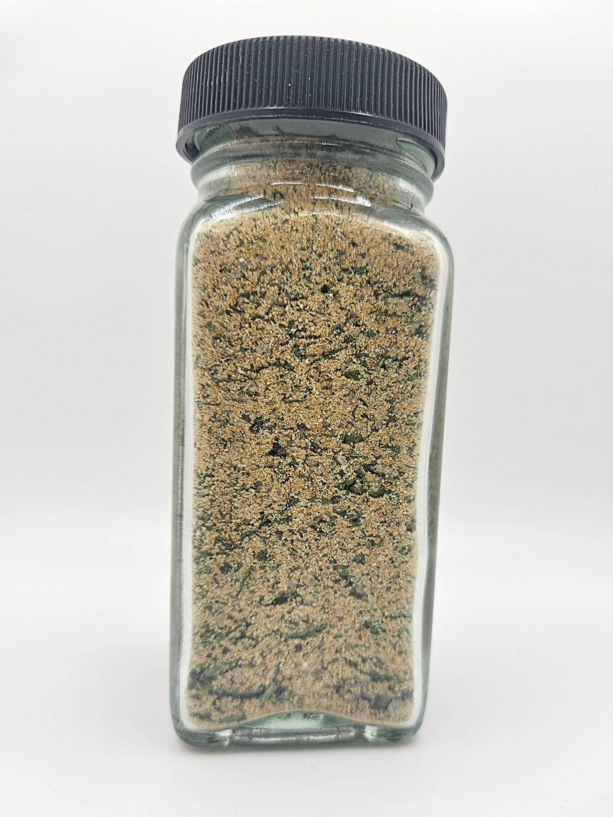 A small glass jar with a black lid containing NEW Pierogi Salt!, a tan seasoning blend with green flecks, ideal for enhancing dishes like pierogi and potatoes.