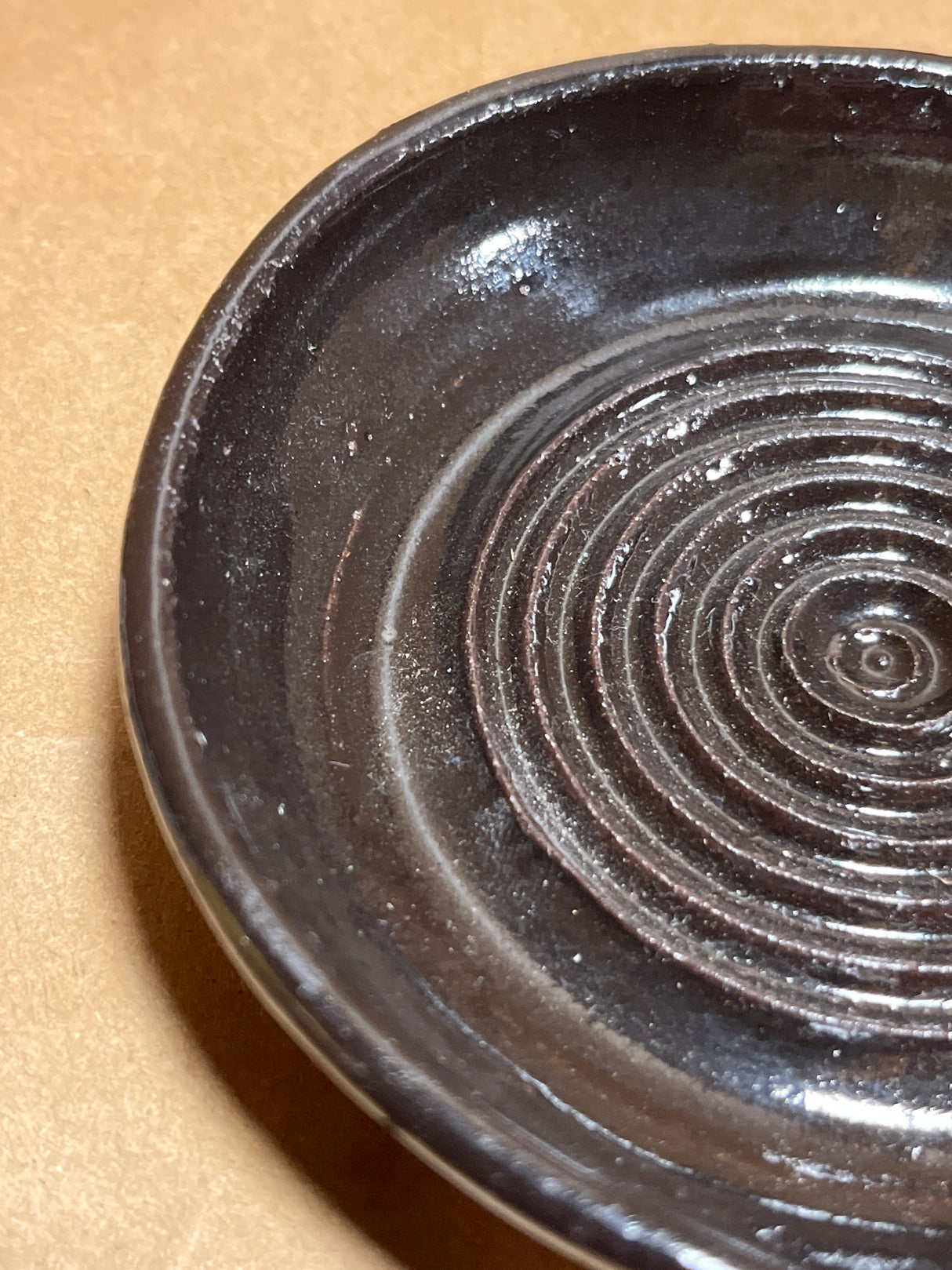 Handmade ceramic garlic grater spoon rest by Charles Lehman, featuring a small, dark-brown glazed dish with concentric ridges, showcased on a neutral surface.