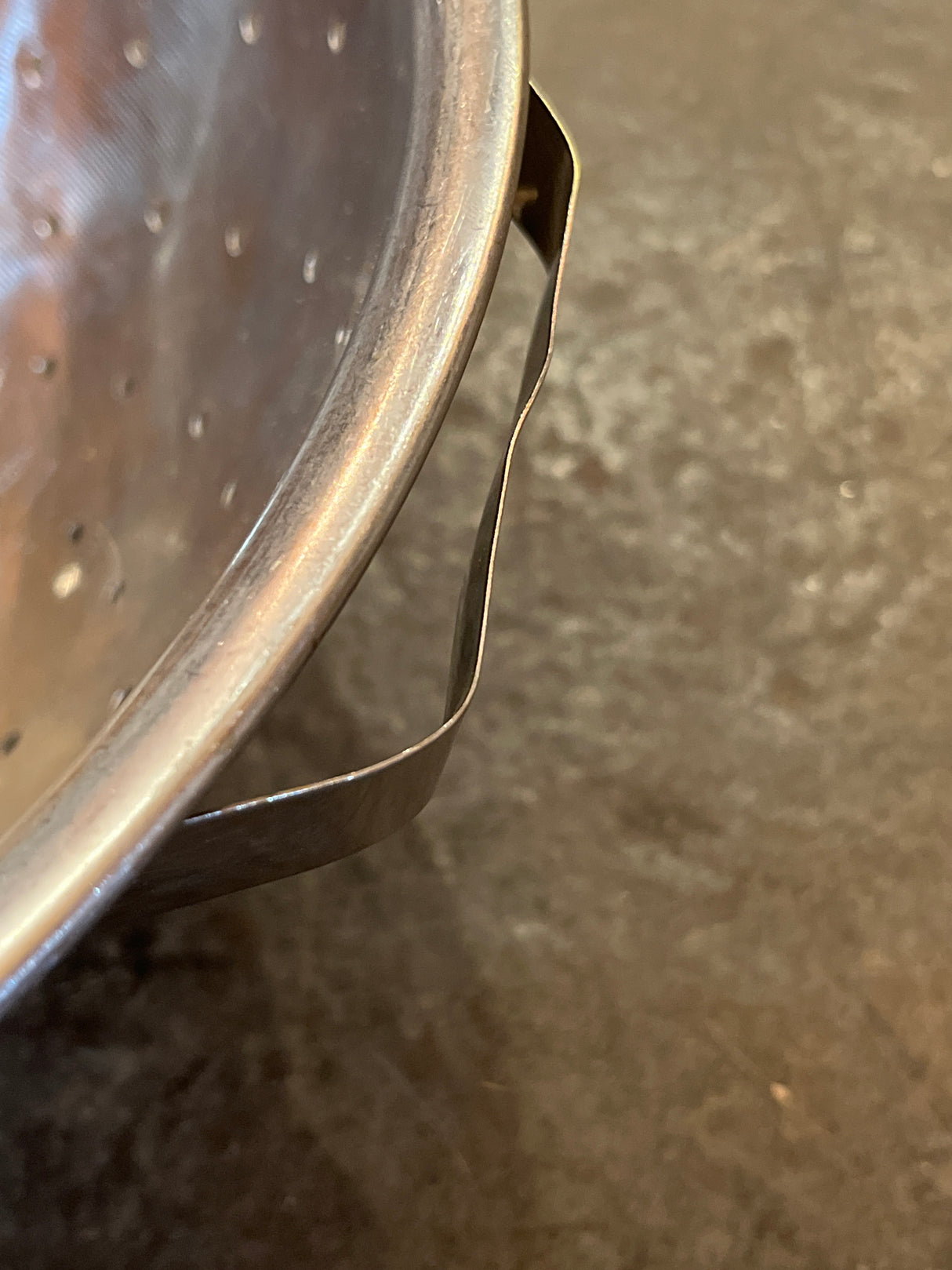 Close-up of Colander with flat base, showing perforated stainless-steel bowl, wire handle, metal clip for hooking over sinks or pots. Handles are thin, slightly bendable.