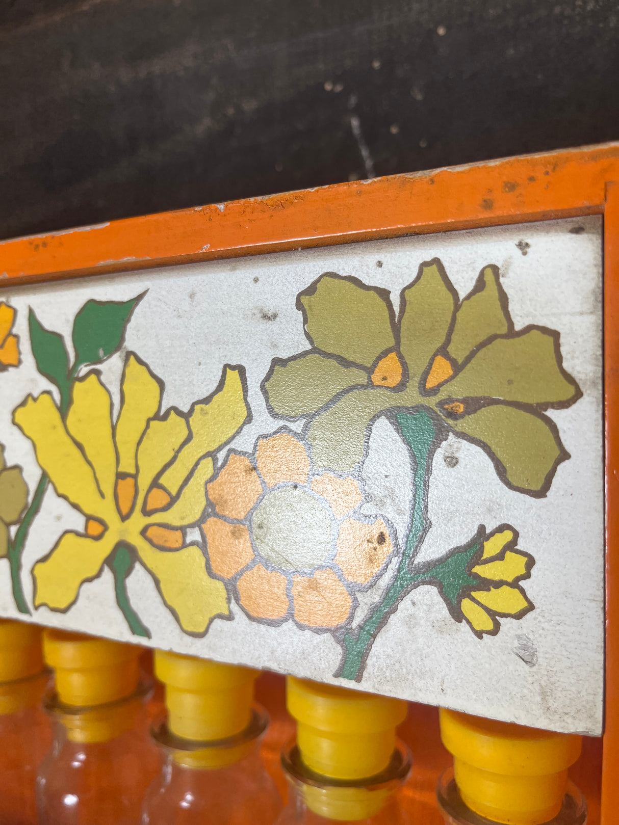 Funky floral wooden spice rack featuring six small glass bottles with yellow lids, complemented by a hand-painted floral motif on the metal panel above.