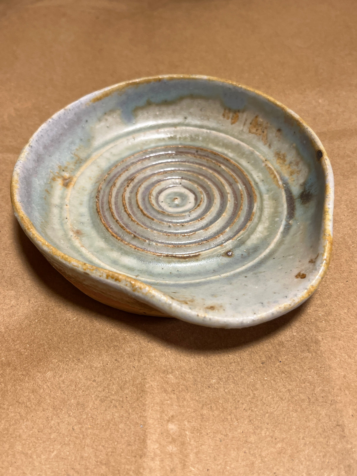 Handmade ceramic garlic grater spoon rest by Charles Lehman, featuring a speckled glaze and concentric rings, displayed on a kraft-paper surface.