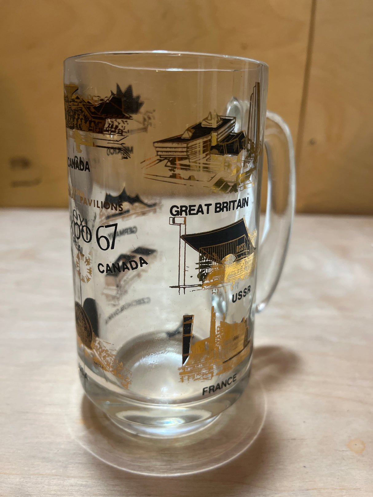 67 Montreal Expo with National Pavilions souvenir mug, a clear glass stein featuring black-and-gold line drawings and labels of various national pavilions, on a wood surface.