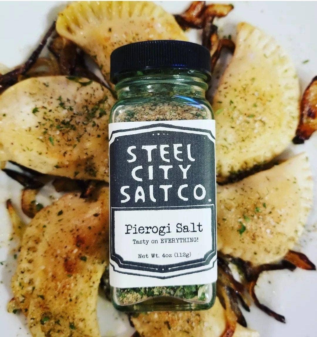 Small glass jar of NEW Pierogi Salt! on a plate of pierogi with caramelized onions and herbs, enhancing this classic dish with its aromatic, flavorful blend.