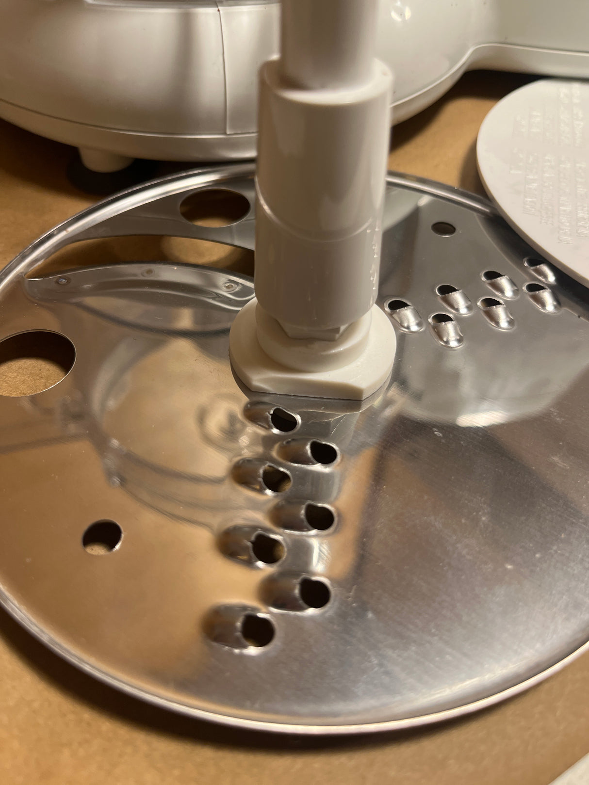 Close-up of the Hamilton Beach Chef Prep 70700 food processor's stainless-steel slicing/grating disc, showing the plastic hub and shaped holes for slicing and shredding.