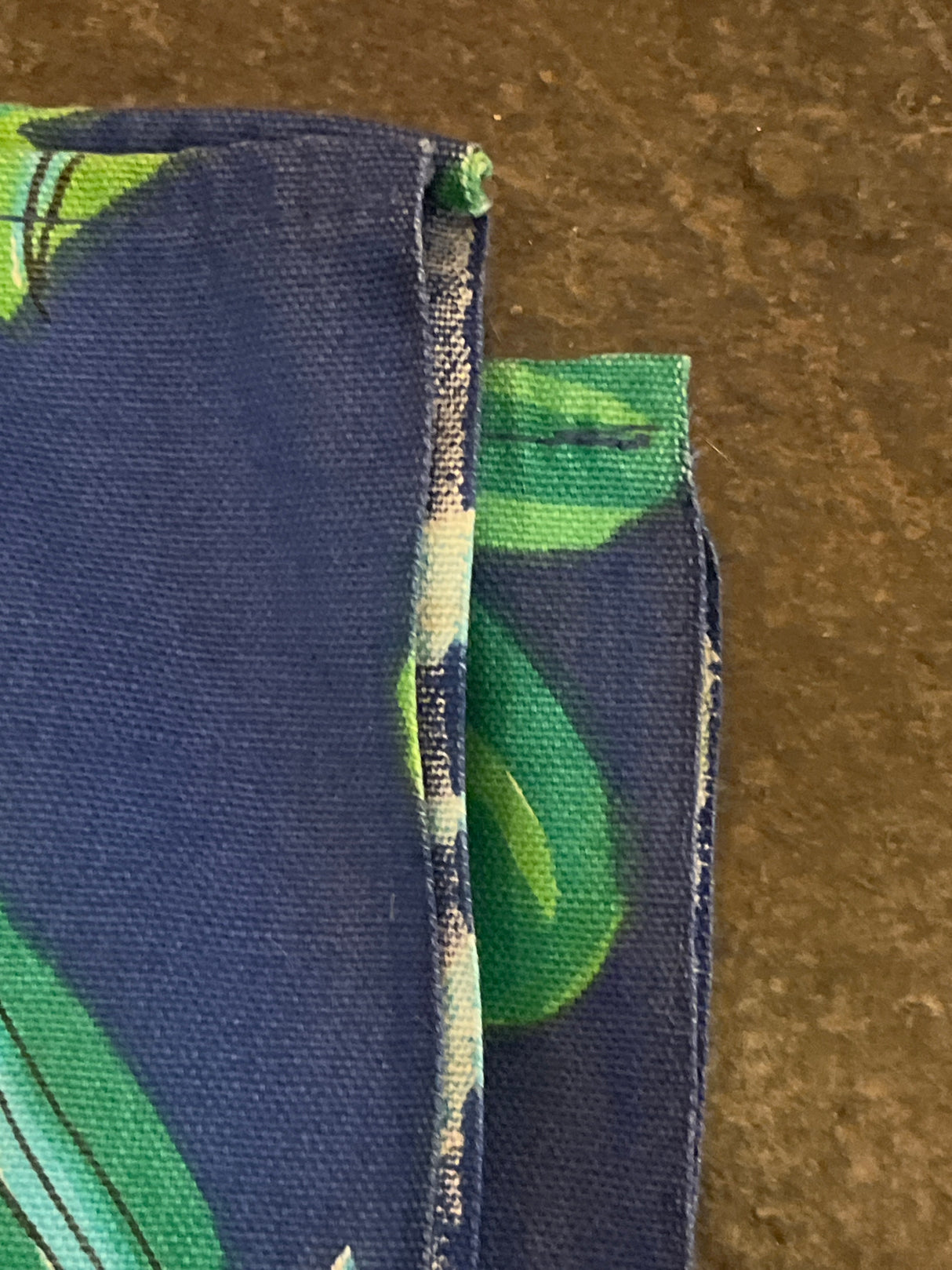 Close-up of a Vintage tablecloth featuring a leafy green pattern on blue fabric, showcasing raw edges and hemmed seams against a stone-like surface.