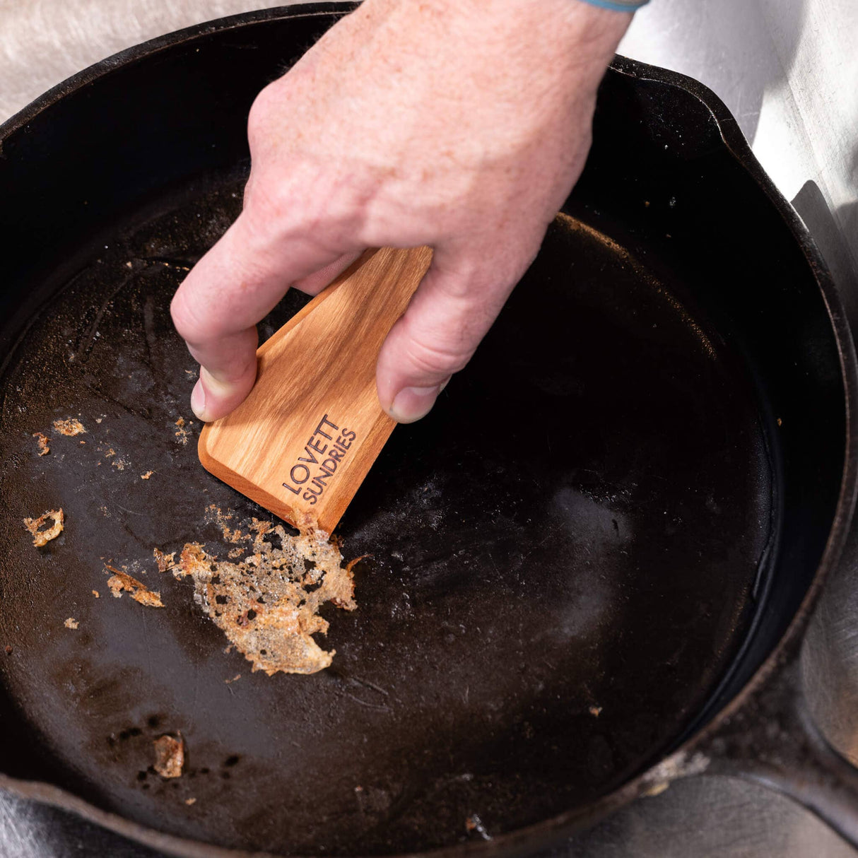 A hand uses the Lovett Sundries Hard Wood Cast Iron Pan Scraper to remove residue from a blackened cast-iron skillet.