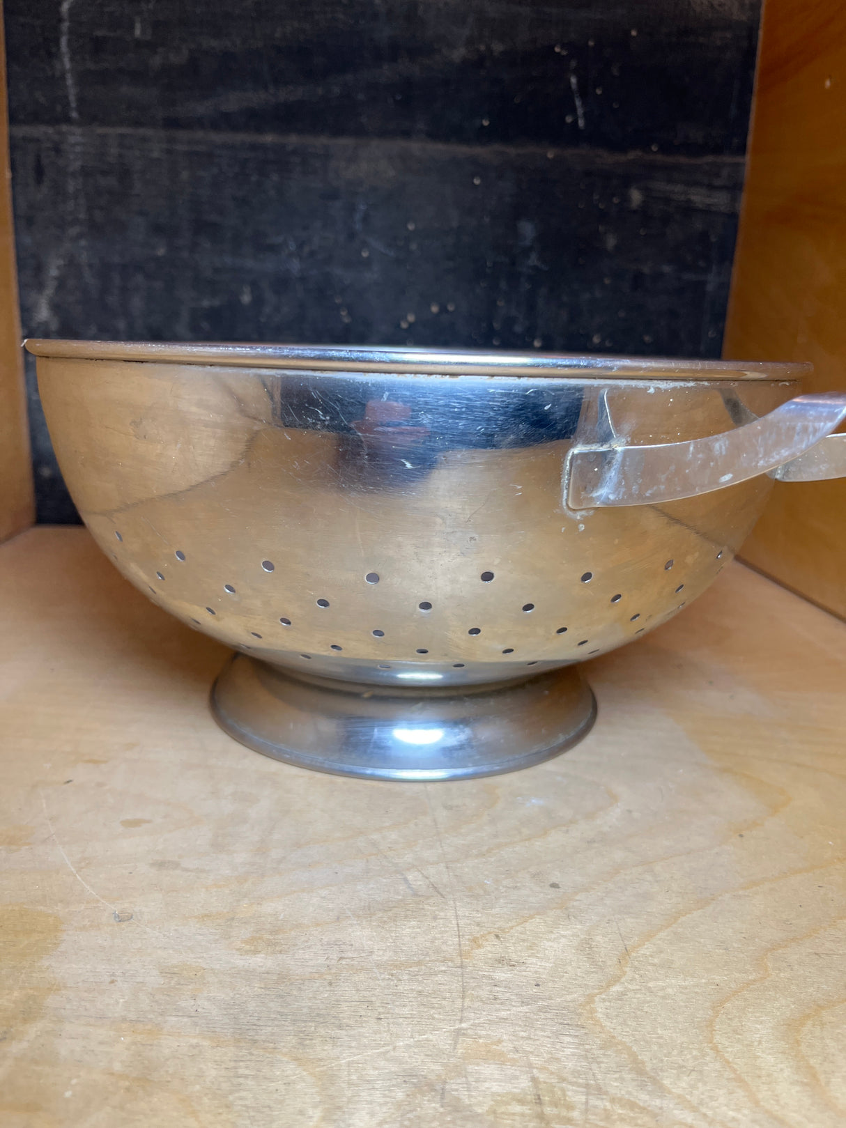 Large colander with handles and elevated base - stainless steel - Sharda, displayed on a wooden shelf, highlighting its sturdy handles and perforated design for effective draining.