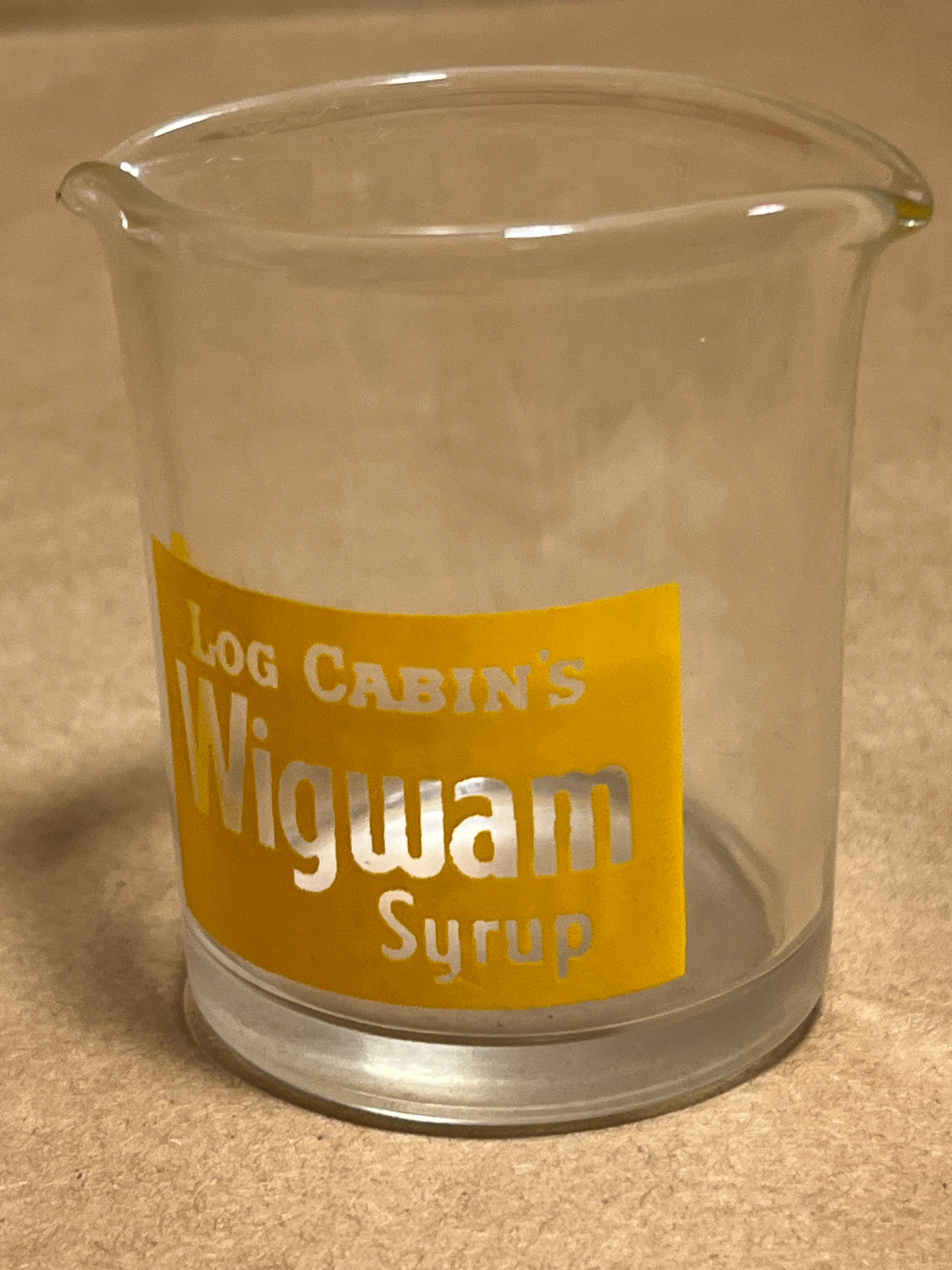 Log Cabin Wigwam syrup pitcher, clear glass with a spout, features a yellow label reading “Log Cabin’s Wigwam Syrup,” designed for easy pouring.