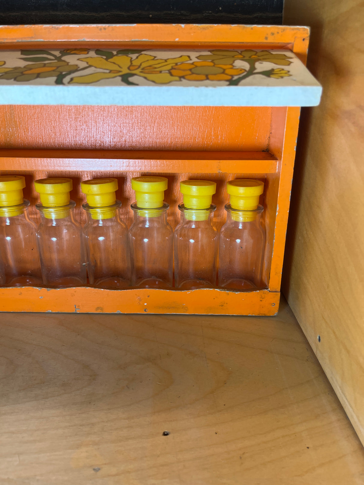 Funky floral wooden spice rack holding six clear glass bottles with yellow stoppers, featuring a hinged shelf with a floral motif, ideal for countertops.