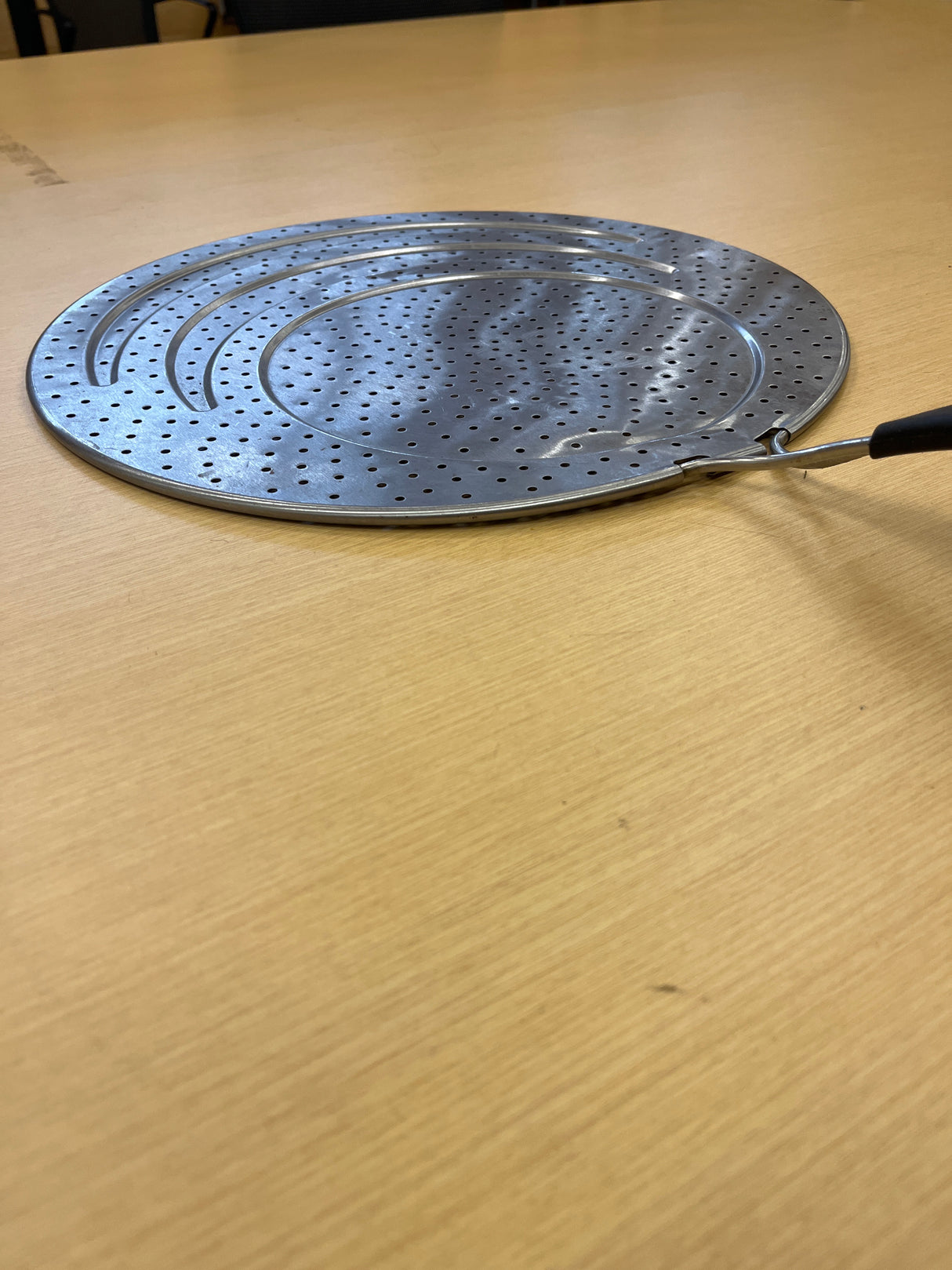 Close-up of Calphalon 13 stainless steel splatter guard with silicone lined handle, designed to fit various pan sizes, placed on a wooden surface.