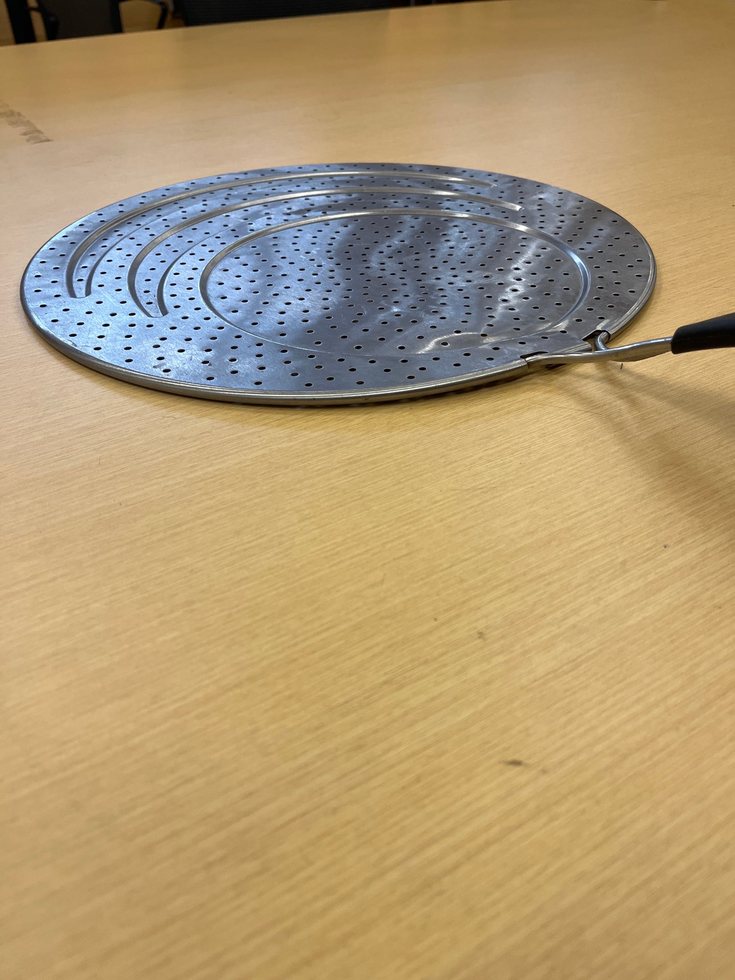 Close-up of Calphalon 13 stainless steel splatter guard with silicone lined handle, designed to fit various pan sizes, placed on a wooden surface.