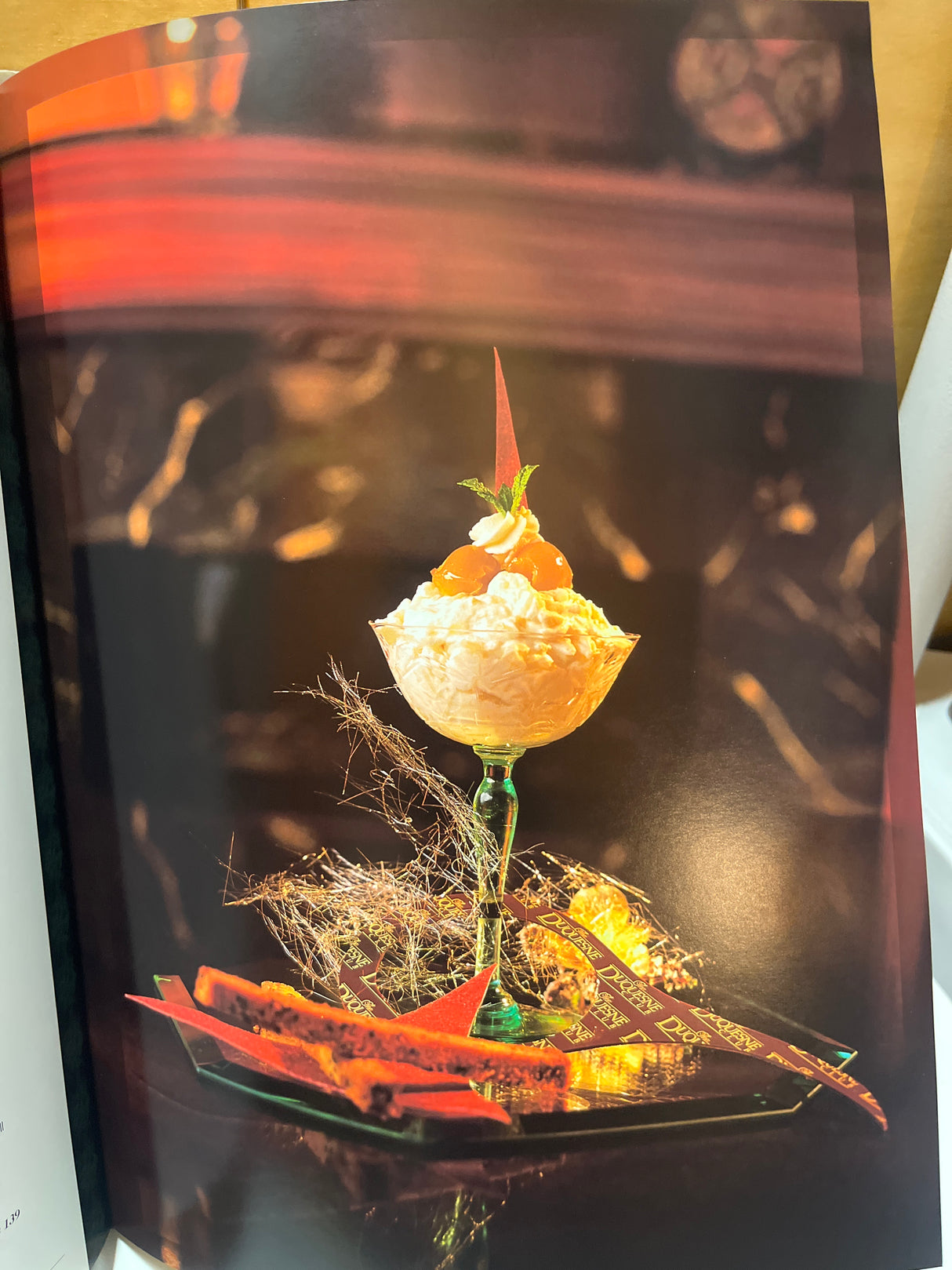 RARE Duquesne Club Cookbook: Artful dessert with fluffy cream in green glass coupe, topped with fruit, mint, and red sugar shard on a dark tray.