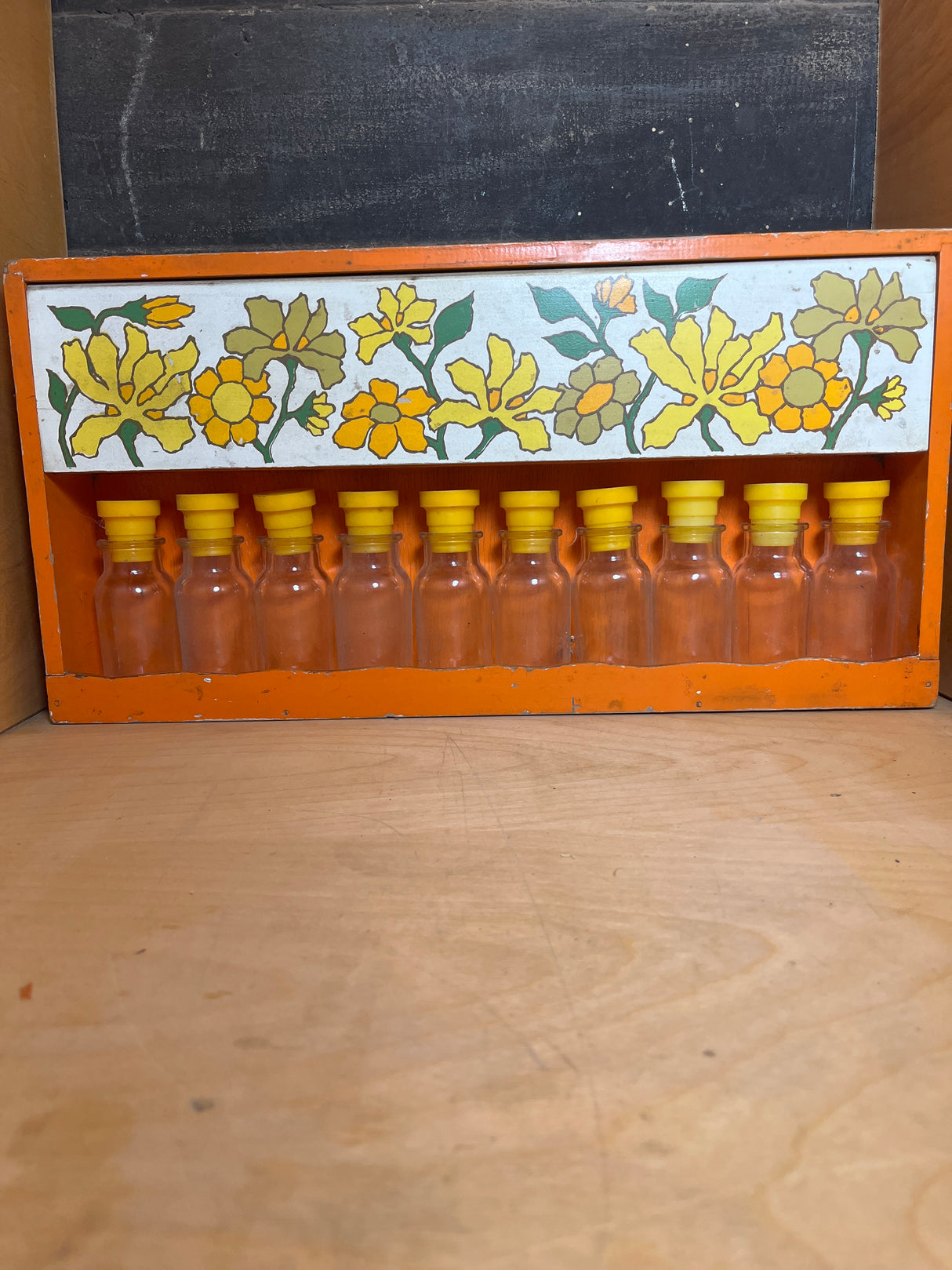 Funky floral wooden spice rack painted orange, featuring a hand-painted floral band and holding 10 clear glass jars with yellow stoppers on the bottom shelf.