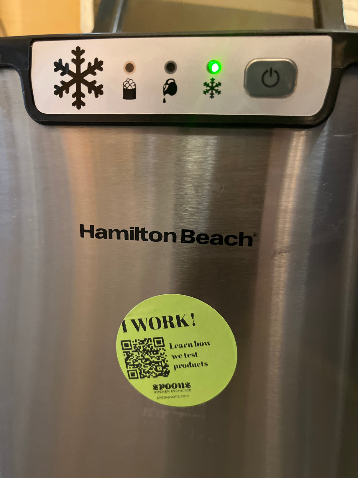 Hamilton Beach portable countertop ice maker with control panel and green snowflake indicator lit, featuring a “Spoons Kitchen Exchange” sticker stating “I WORK!” with a QR code.