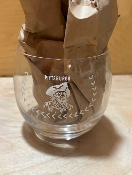 RARE 1960 Pittsburgh Pirates World Champions commemorative drinking glass featuring a tricorn-hat frontiersman graphic, stuffed with crumpled brown paper, on a wooden surface.