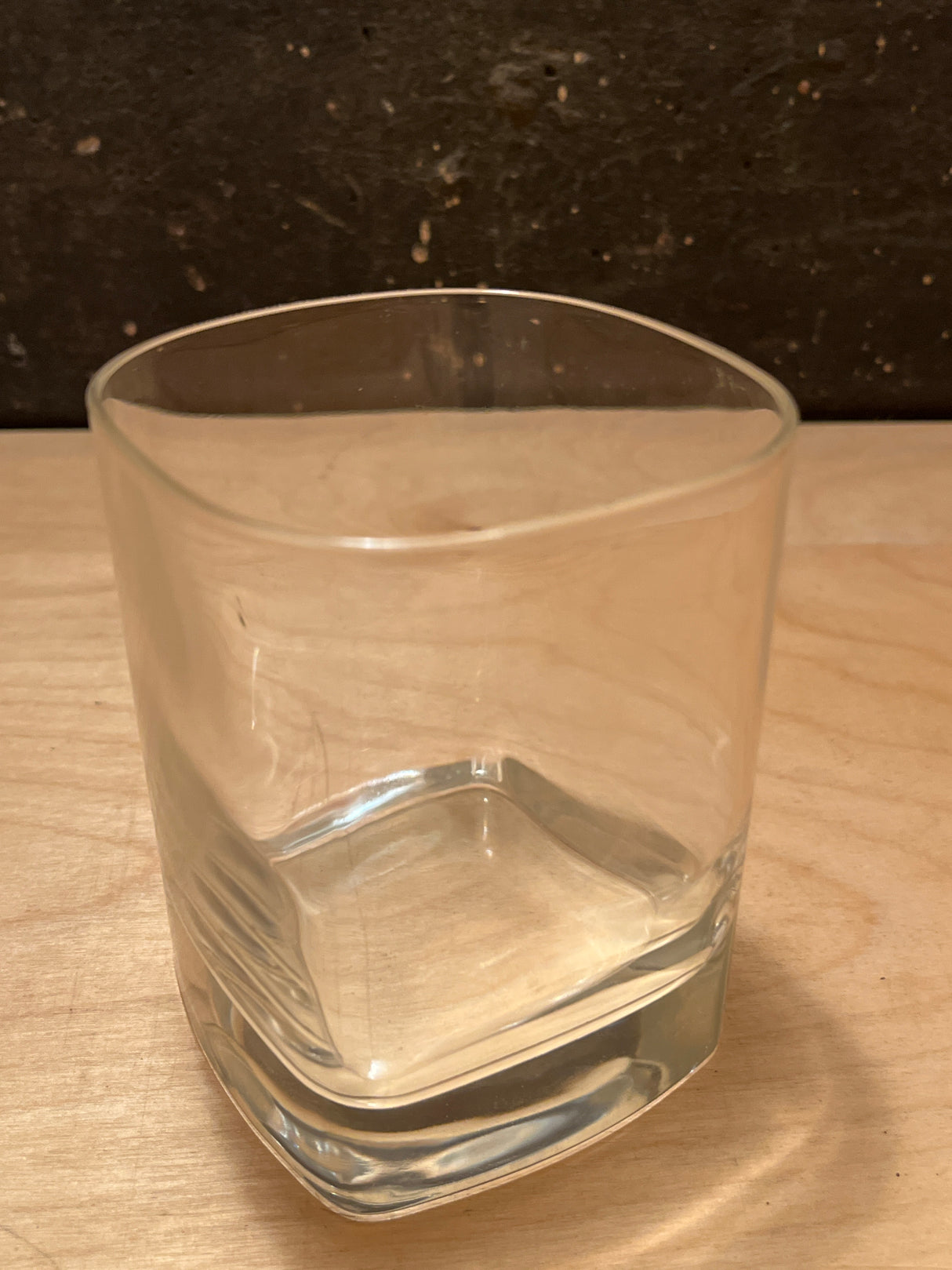 Square heavy bottom tumbler drinking glass with gently rounded square sides, sitting on a light wood surface, perfect for serving drinks with elegance and stability.