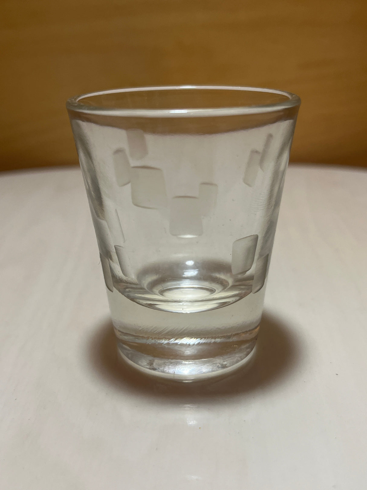 Anchor Hocking cubist shot glass featuring an etched pattern of overlapping rectangles, partially filled with liquid, on a light tabletop with a wooden backdrop.
