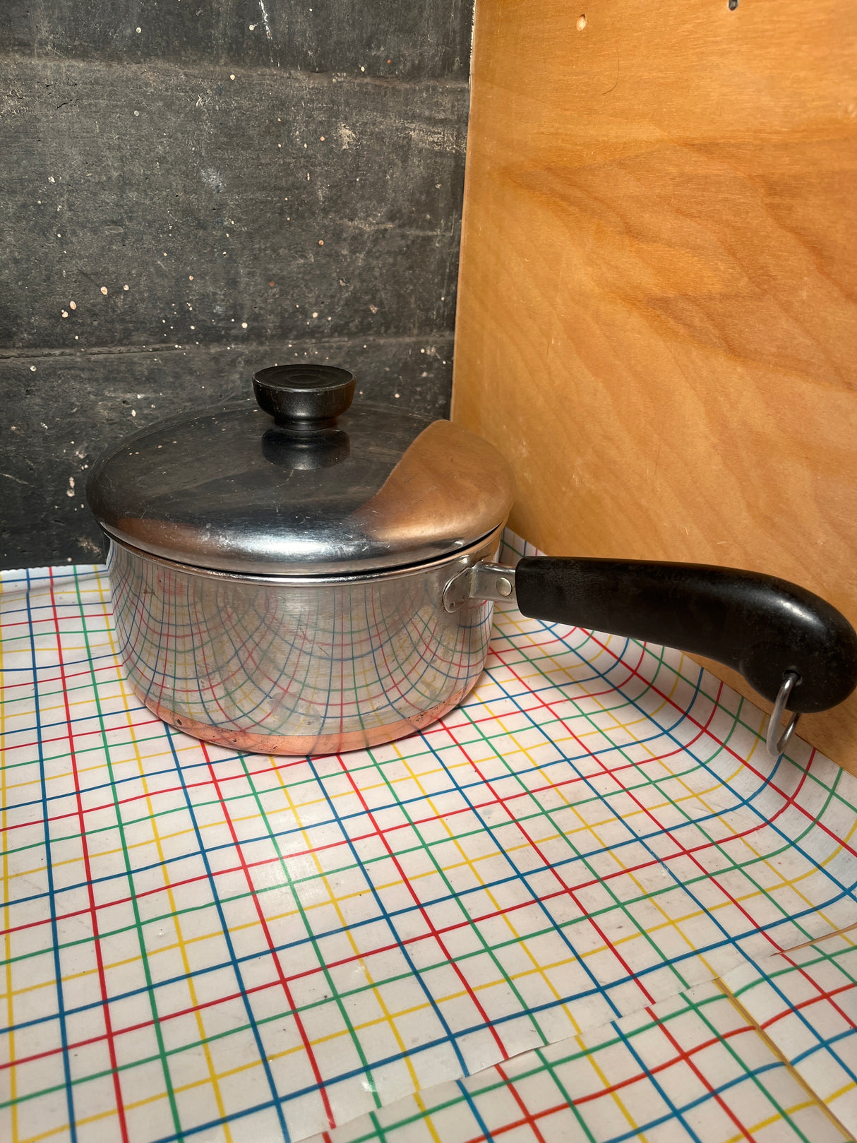 2-qt saucepan with copper bottom - vintage Revere Ware stainless steel, featuring a black plastic handle, sits on a multicolored grid-patterned table covering.