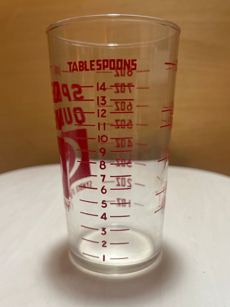 Speed Queen glass measuring cup, empty, shows red gradations for tablespoons and ounce markings, placed on a tabletop with a wood-tone background.