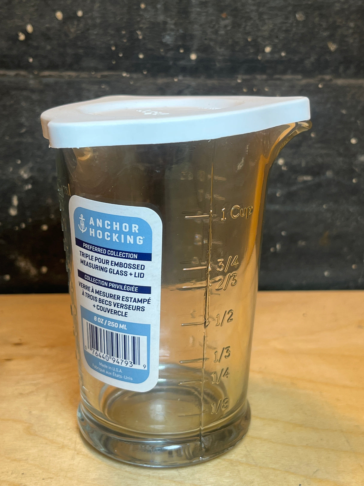Triple pour embossed measuring glass with silicone lid by Anchor Hocking, featuring embossed volume markings and a white plastic lid, displayed on a wooden surface.