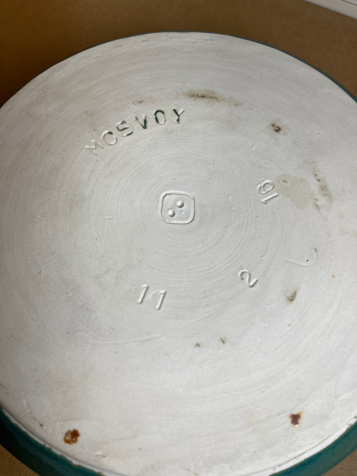 Unglazed underside of McEvoy ceramic dip dish, showing white clay with wheel-throw lines, maker’s mark, stamped “MOSEVOY,” numerals “10,” “2,” “11,” and hint of teal glaze.