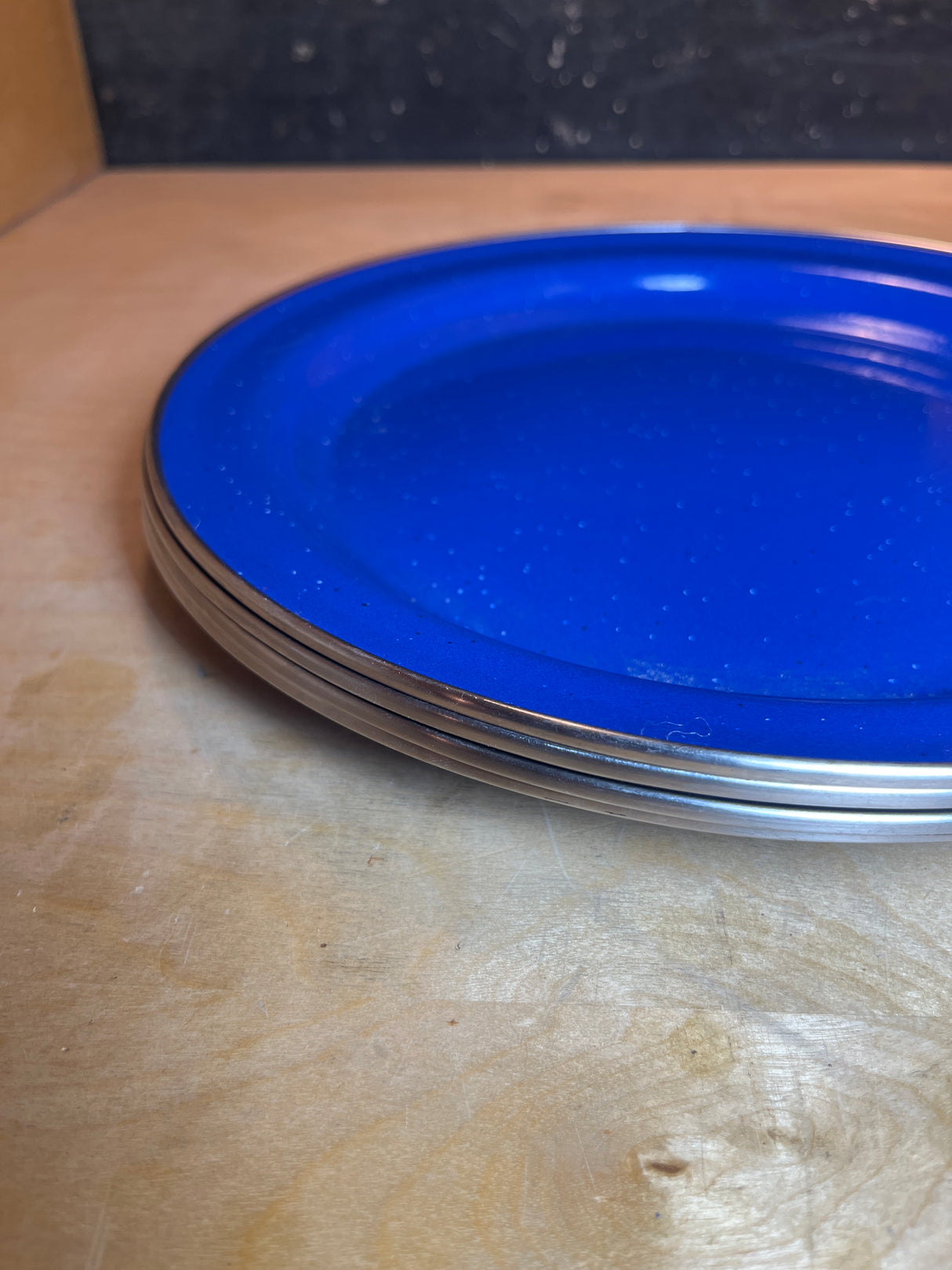 Set of 4 blue speckled enamelware plates with metal rims, stacked on a light wood shelf, ideal for camping and rustic dining.