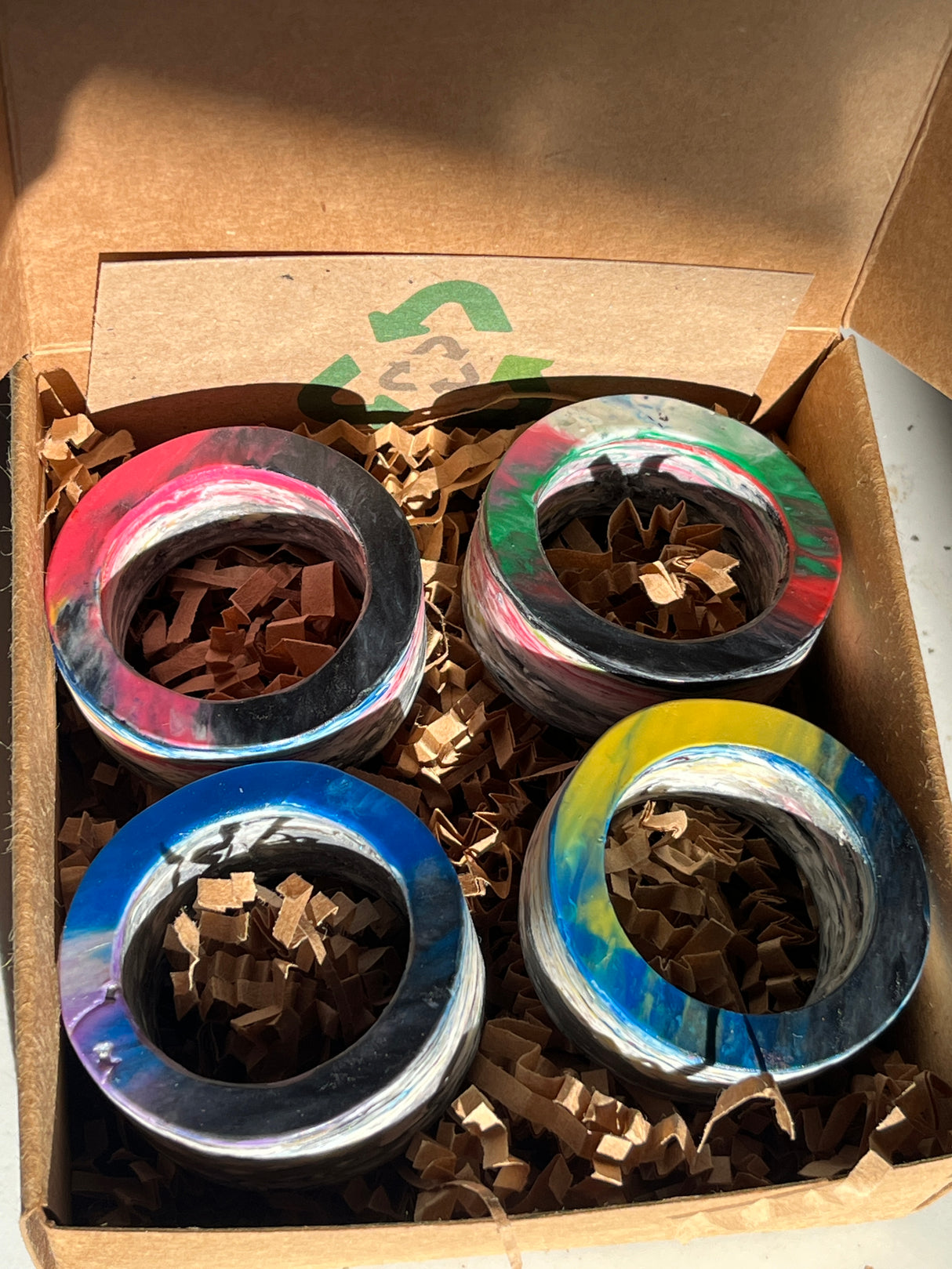 Napkin rings from Reimagined Recycling, crafted from swirled resin, featuring varied patterns. Packed in a box with crinkled paper for protection. Unique and eco-friendly design.