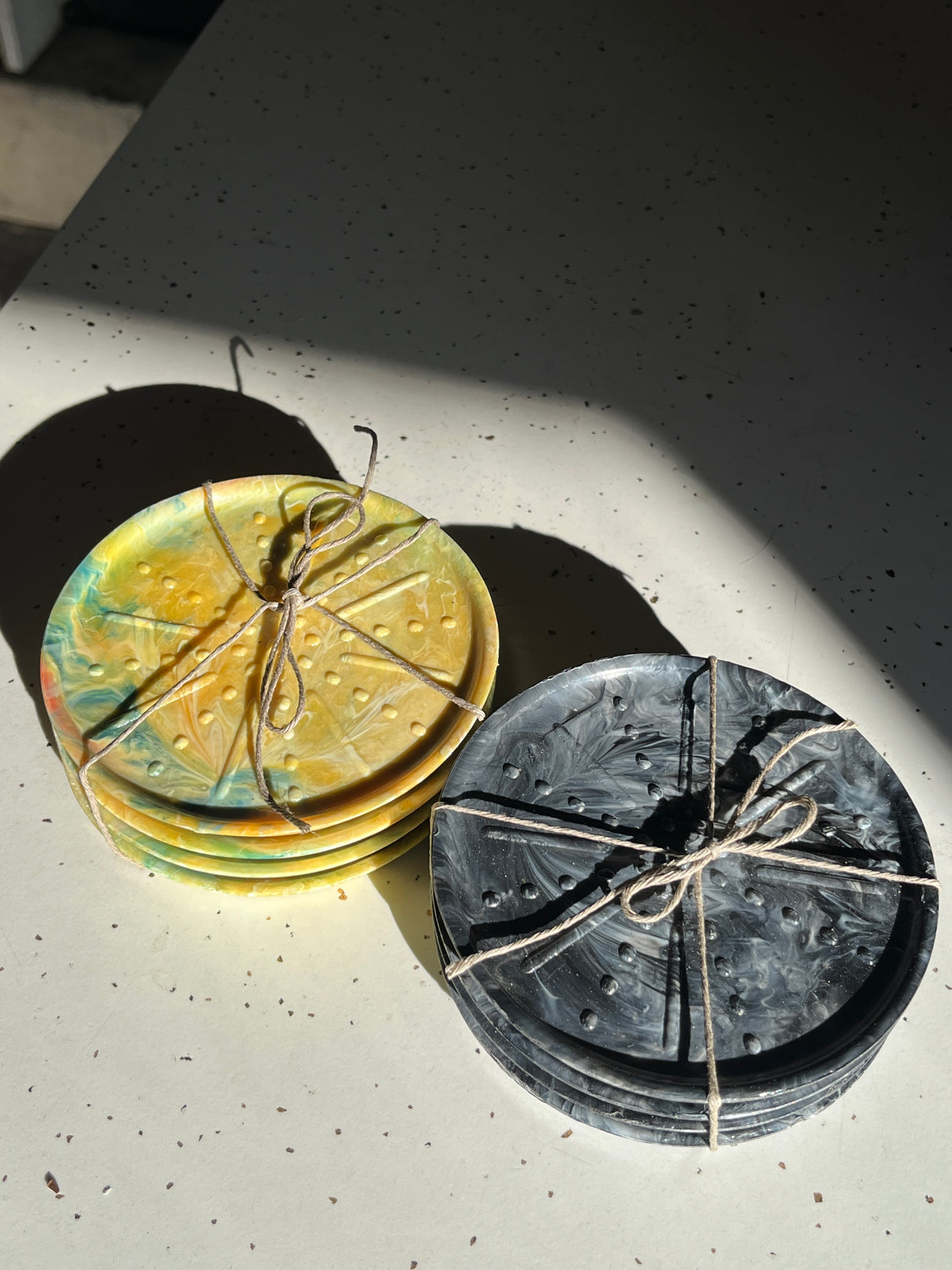 Coaster Sets from Reimagined Recycling: Two stacks of hand-glazed ceramic coasters with drainage dimples, tied with twine, showcasing marbled and swirl designs.