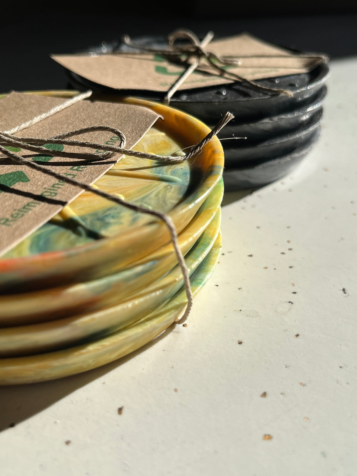 Coaster Sets from Reimagined Recycling: two stacks of round coasters, yellow/green marbled and solid black, wrapped in twine with labels, on a tabletop.