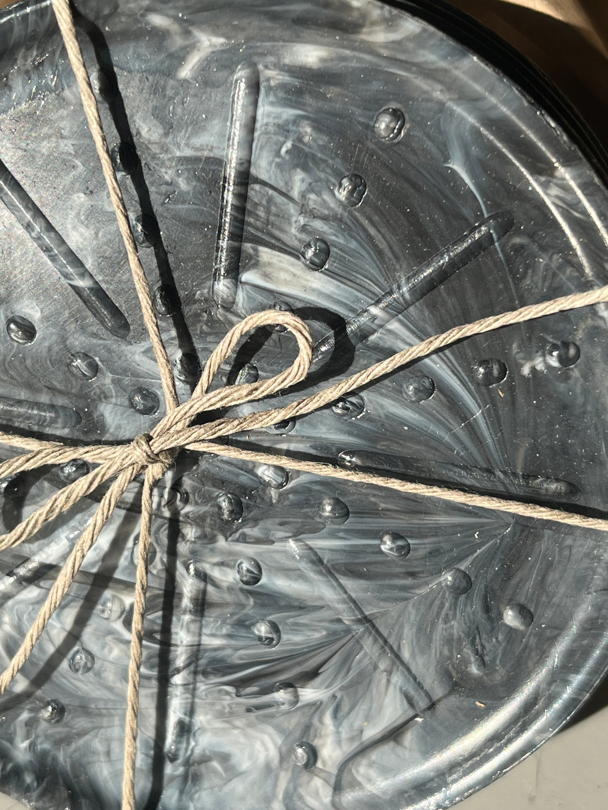 Coaster Sets from Reimagined Recycling feature a blackish-gray swirled surface with raised dots and ribs, tied with rough twine, made from recycled #5 plastics.