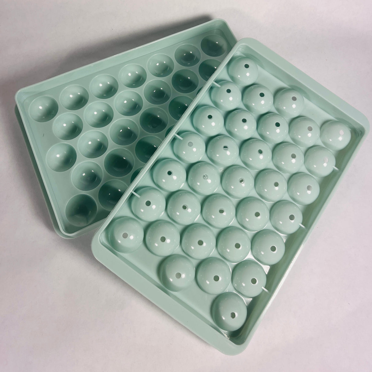 Turquoise Wibimen mini spherical ice tray, with the top lid positioned at an angle so you can see the inside of the other tray. Set on a white background.