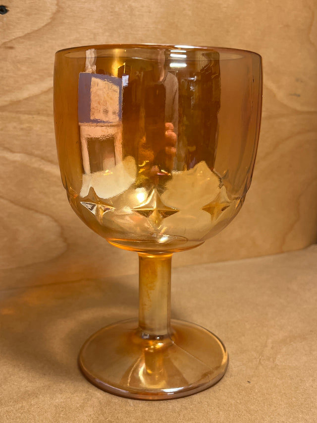 Bartlett Collins Star Marigold Carnival orange glass water goblet with a short stem and rounded bowl, featuring embossed star or diamond motifs, on a wooden surface.