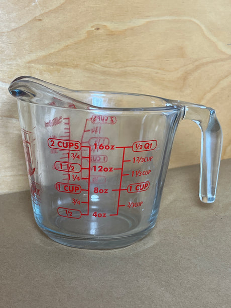 Anchor Hocking glass 2-cup measuring cup with handle, featuring red volume markings and a pouring spout, rests on a light wood surface.