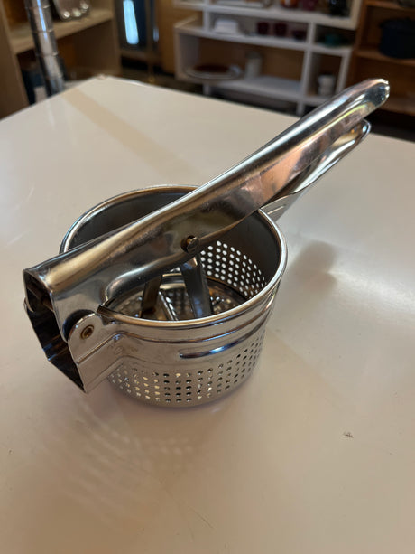 Stainless steel Ricer and Masher for potatoes, fruit, and juice on a white countertop, crafted for durability and smooth purée creation.