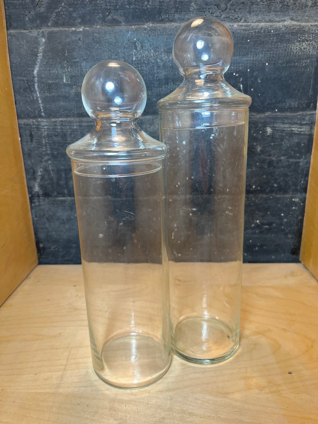 Libbey tall apothecary clear glass jars with lids sit side by side, showcasing their elegant cylindrical shape and round knob-topped lids on a wooden surface.
