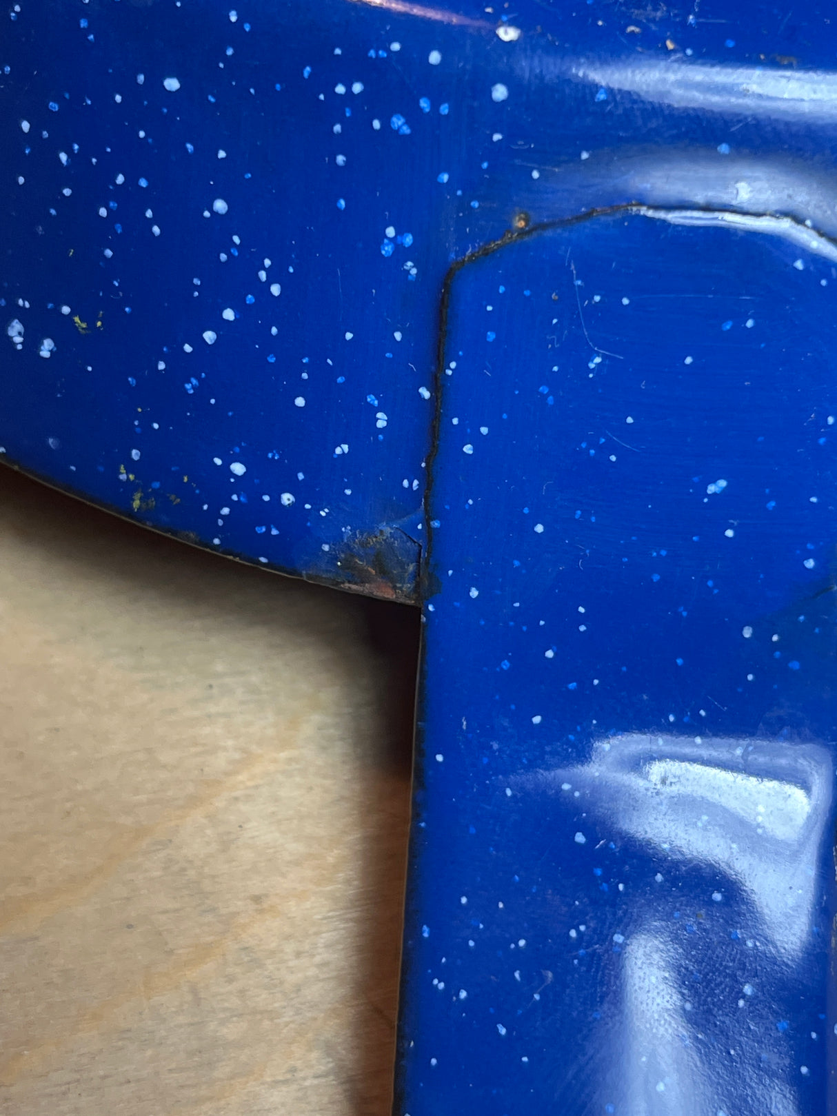 Blue speckled enamelware fry pan for camping on wood surface, showing metal corner seam and minor enamel chipping, ideal for safe outdoor cooking with long handle.