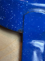 Blue speckled enamelware fry pan for camping on wood surface, showing metal corner seam and minor enamel chipping, ideal for safe outdoor cooking with long handle.