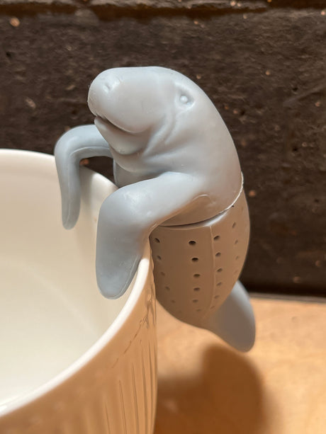ManaTea infuser shaped like a manatee, hooks over a mug's rim. The perforated bottom holds loose-leaf tea, made from food-safe silicone for charming brewing.